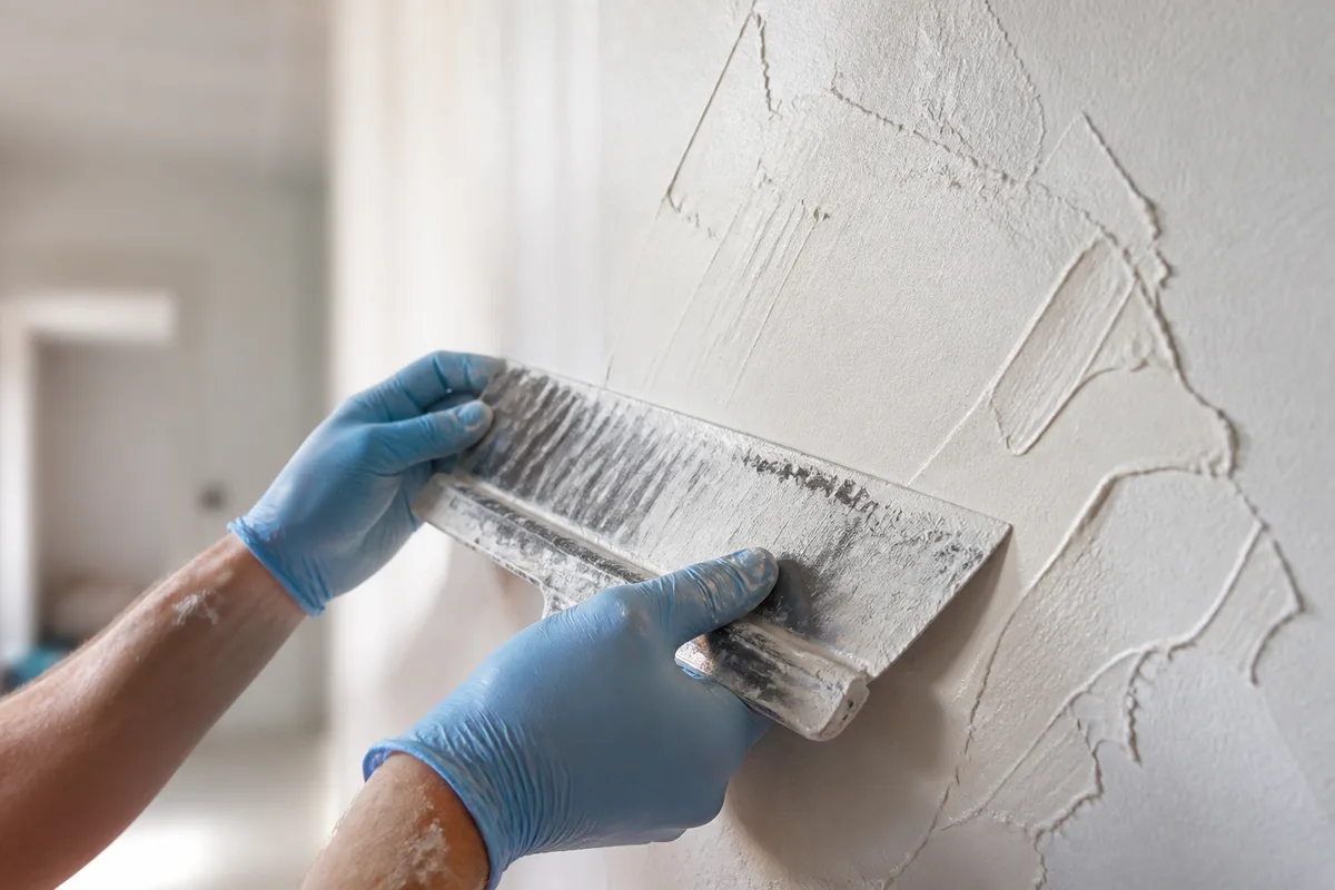 Mains gantées lissant un enduit clair sur un mur d’appartement en travaux